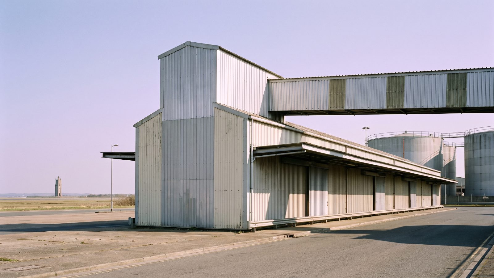A long steel freight handling shed on a port estate with an enclosed conveyor gantry running along it, round storage tanks, flat estuary marshland.