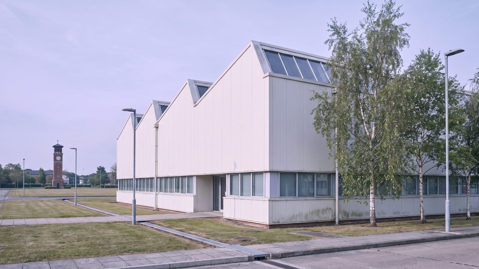 A 1980s manufacturing unit on a landscaped industrial estate, saw-tooth roof with north-light glazing, pale cladding with a band of windows, birch trees in front.