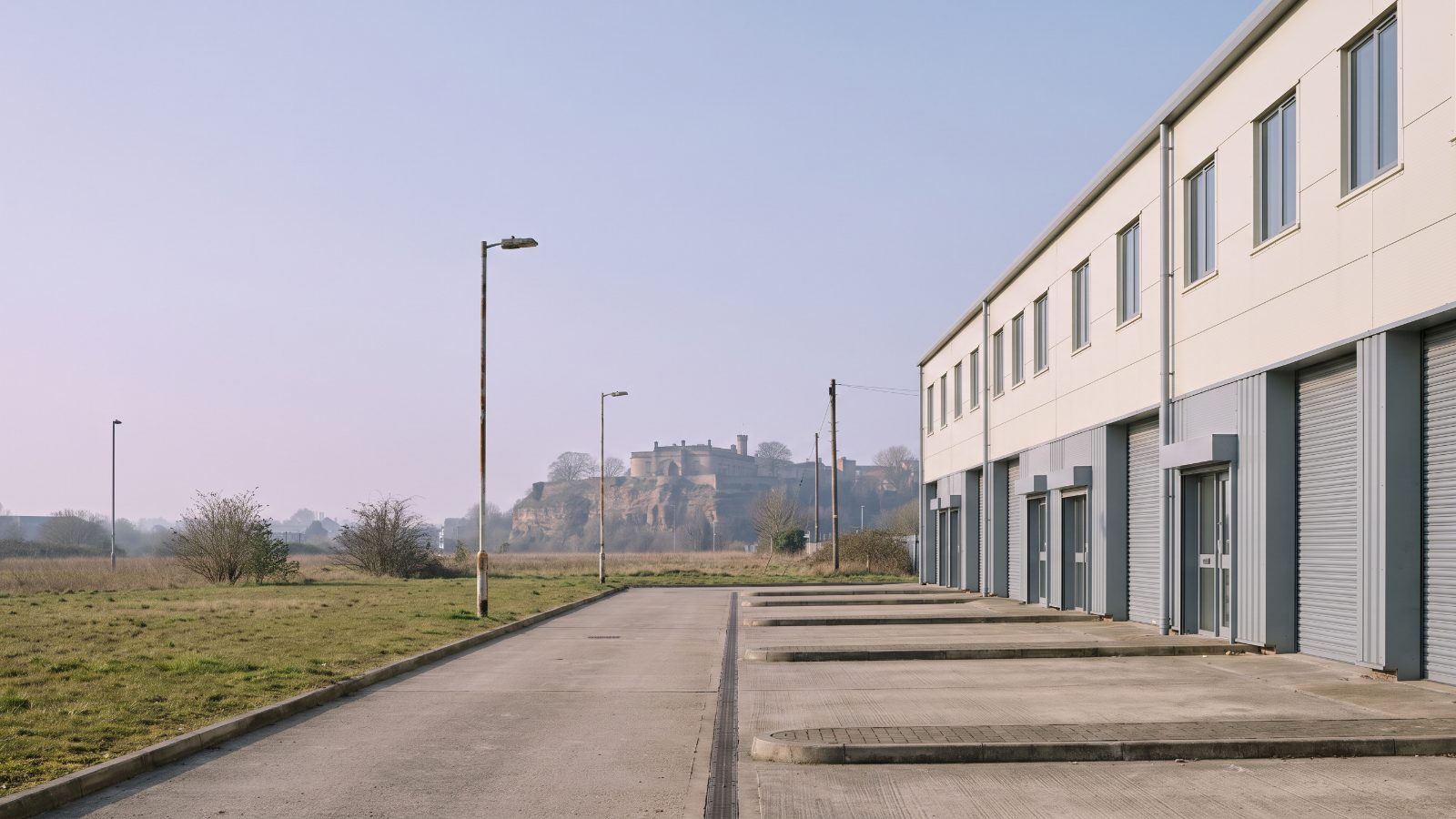 A terrace of small light-industrial units on a tidy trading estate, each with a grey roller shutter and a pale clad upper panel, a concrete apron and low kerbs in front. A castle on a sandstone rock stands in the middle distance.