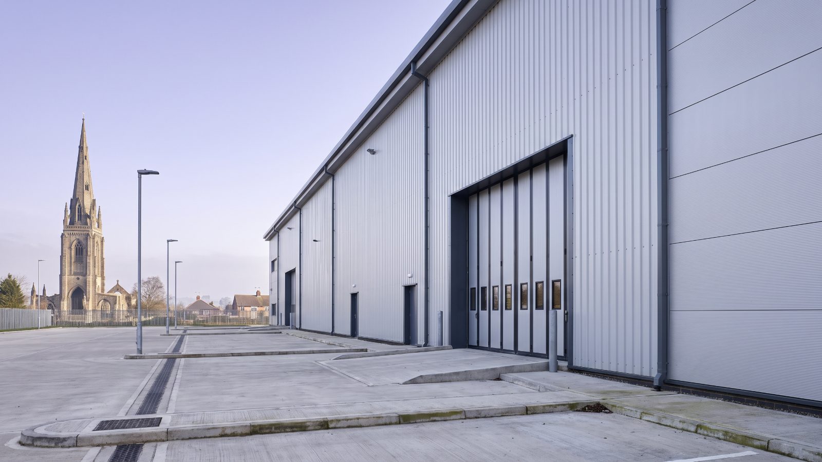 A tall automotive-parts distribution warehouse with pale silver cladding and a high loading door, a clean concrete yard. The tall gothic spire of a ruined cathedral stands on the skyline in the middle distance.