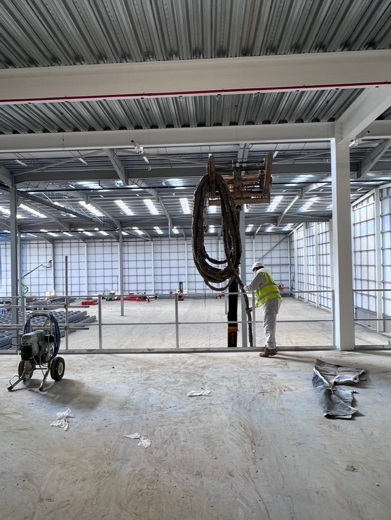 During: a spray rig and its heated hose set up on the concrete deck, an operative in protective clothing feeding the hose over the edge rail, the profiled steel soffit above.