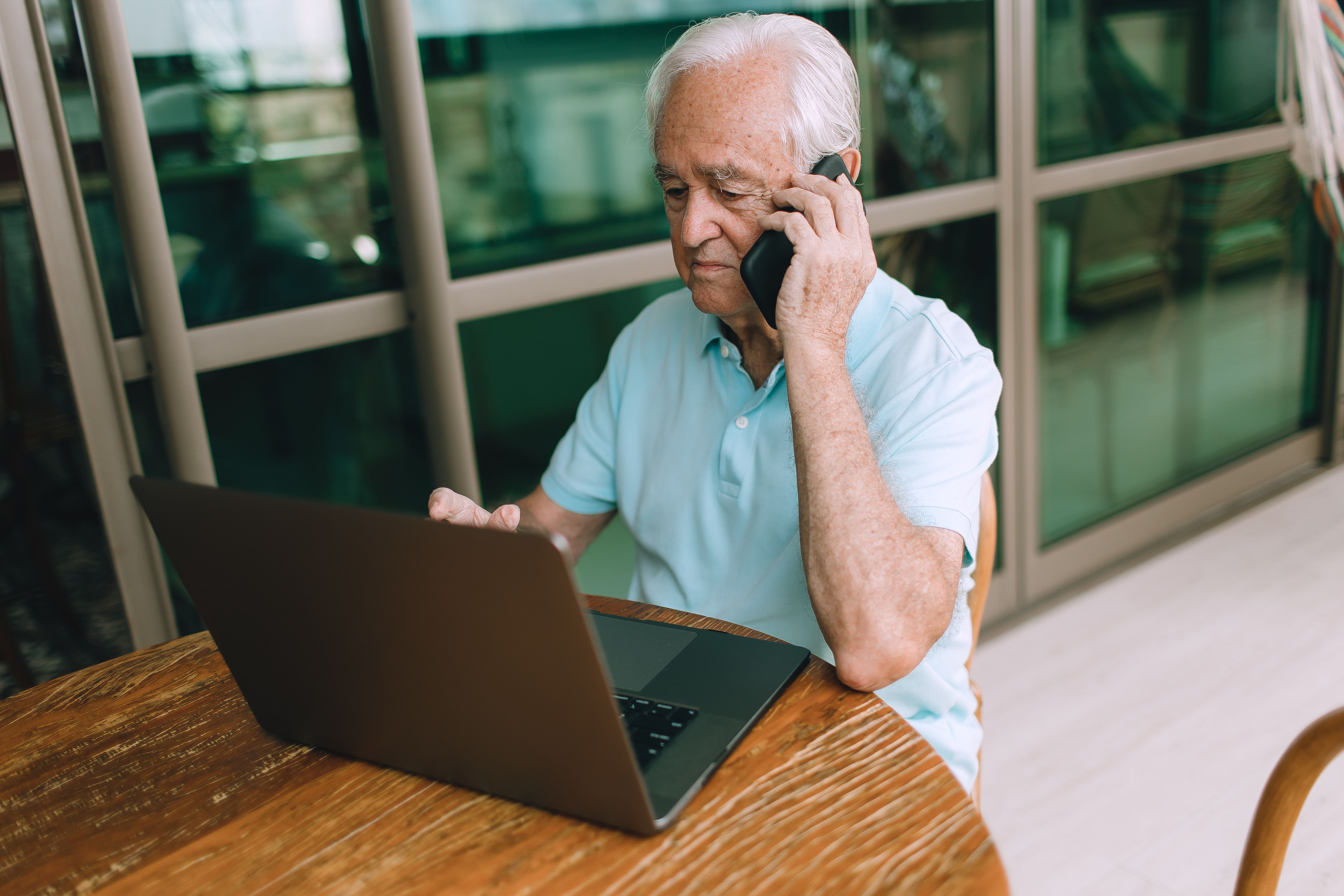 Elderly man in light blue shirt using laptop and talking on smartphone indoors.