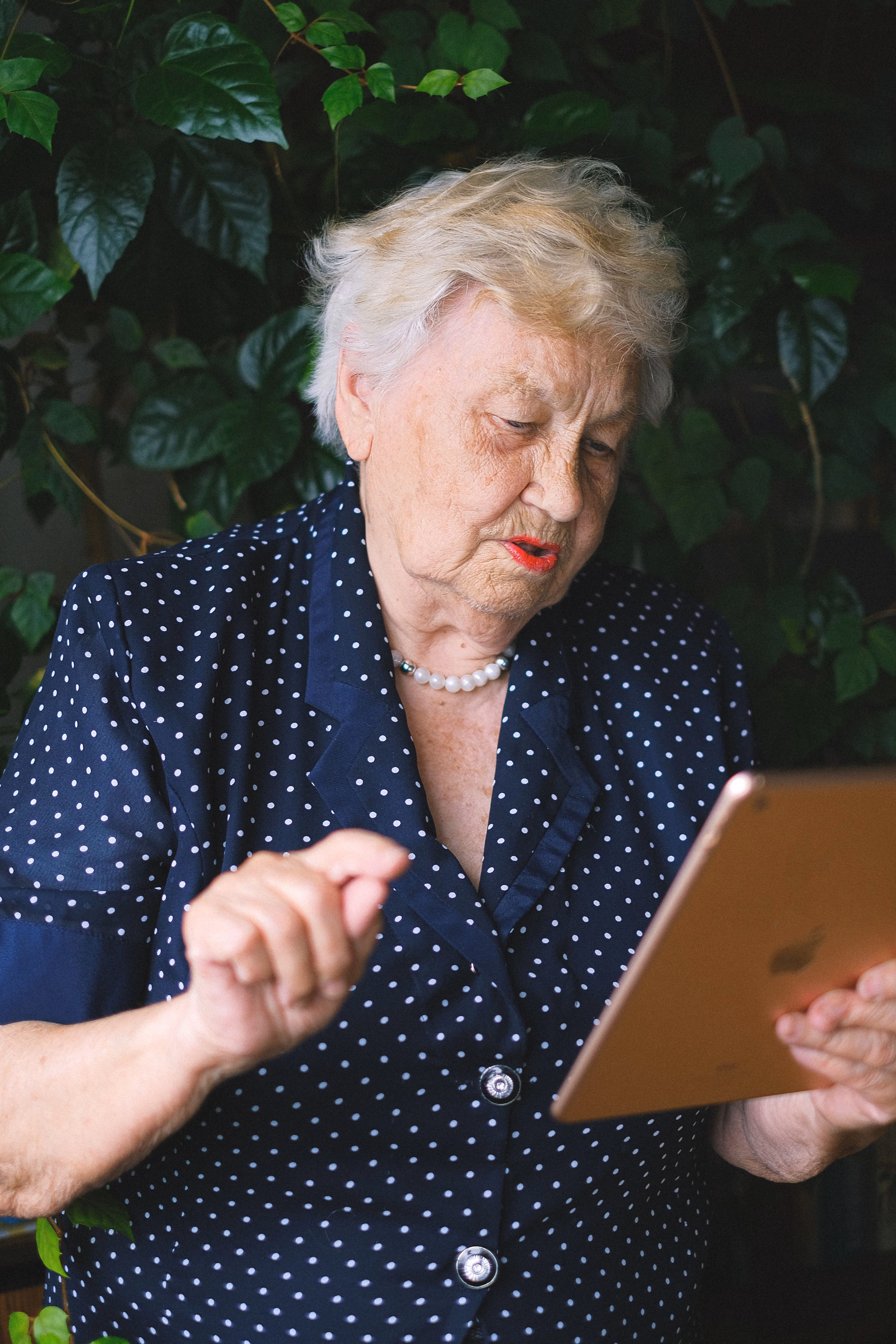 Elderly woman in a navy polka-dot blouse using a tablet with a background of green leaves.