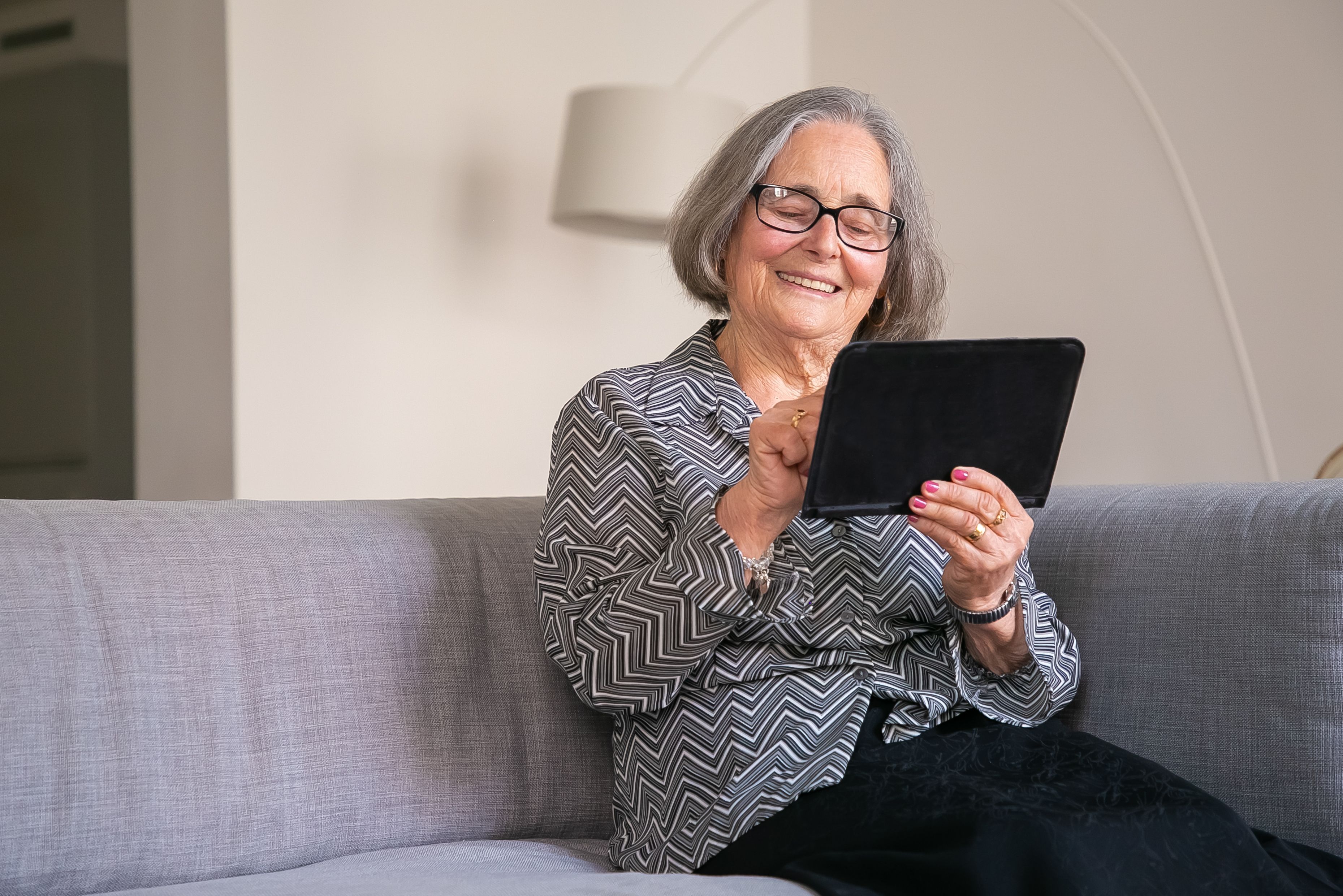 Smiling elderly woman with glasses using a tablet while sitting on a gray couch.