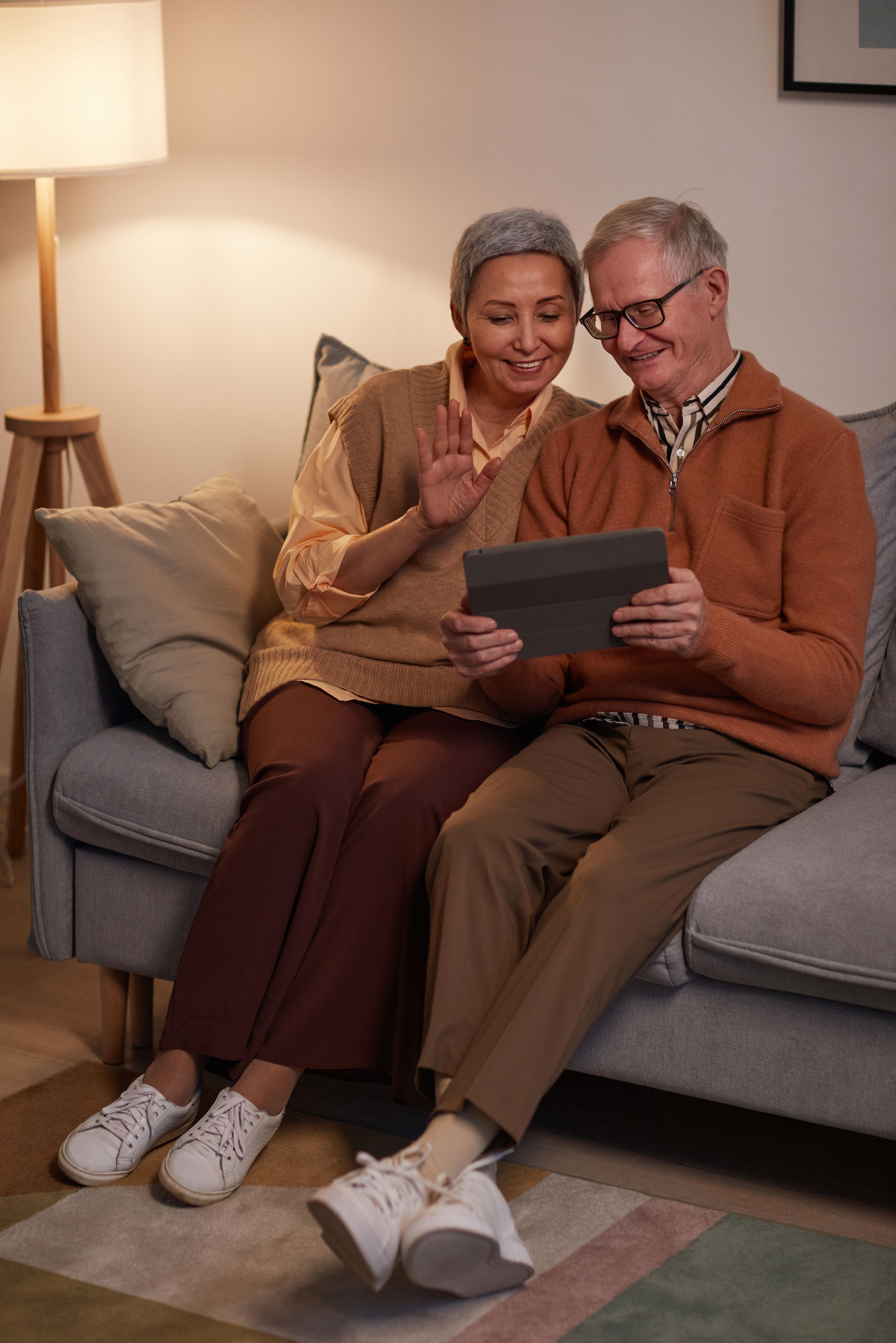 Elderly couple sitting on a couch using a tablet, woman waving and smiling at the screen.