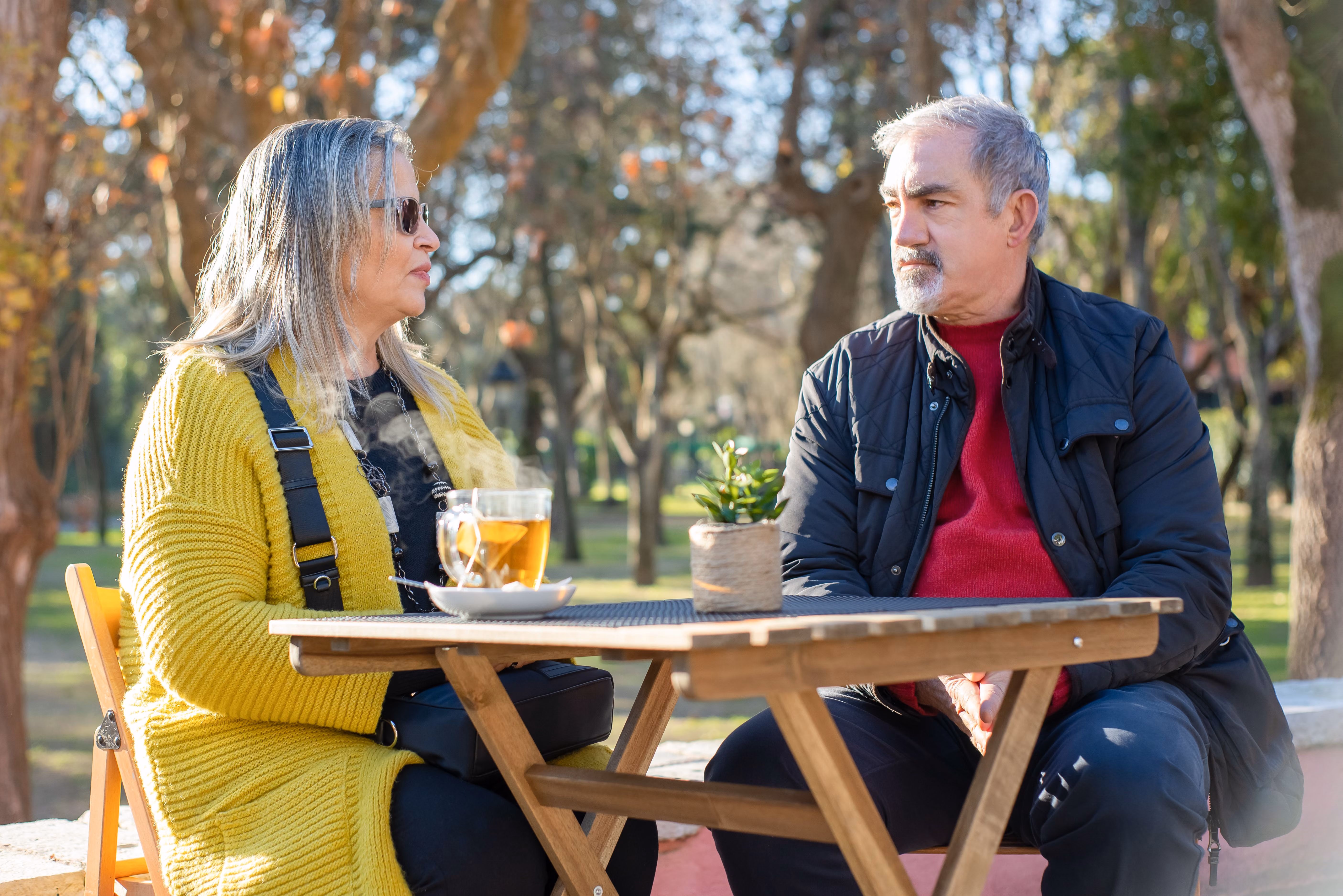 Older man and woman sitting at an outdoor wooden table with a steaming cup of tea between them.
