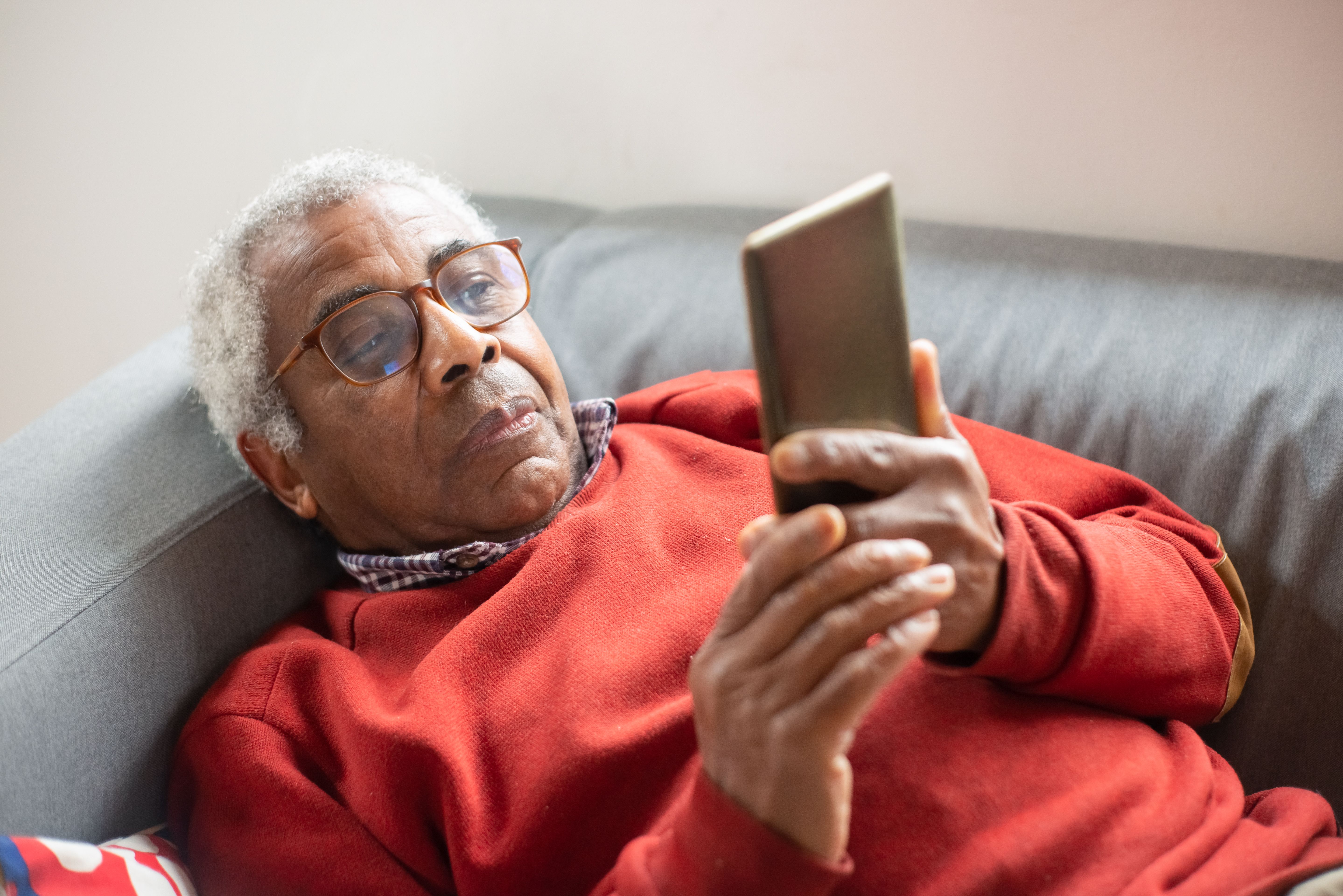 Elderly man in glasses wearing a red sweater lying on couch, looking at a smartphone in his hands.