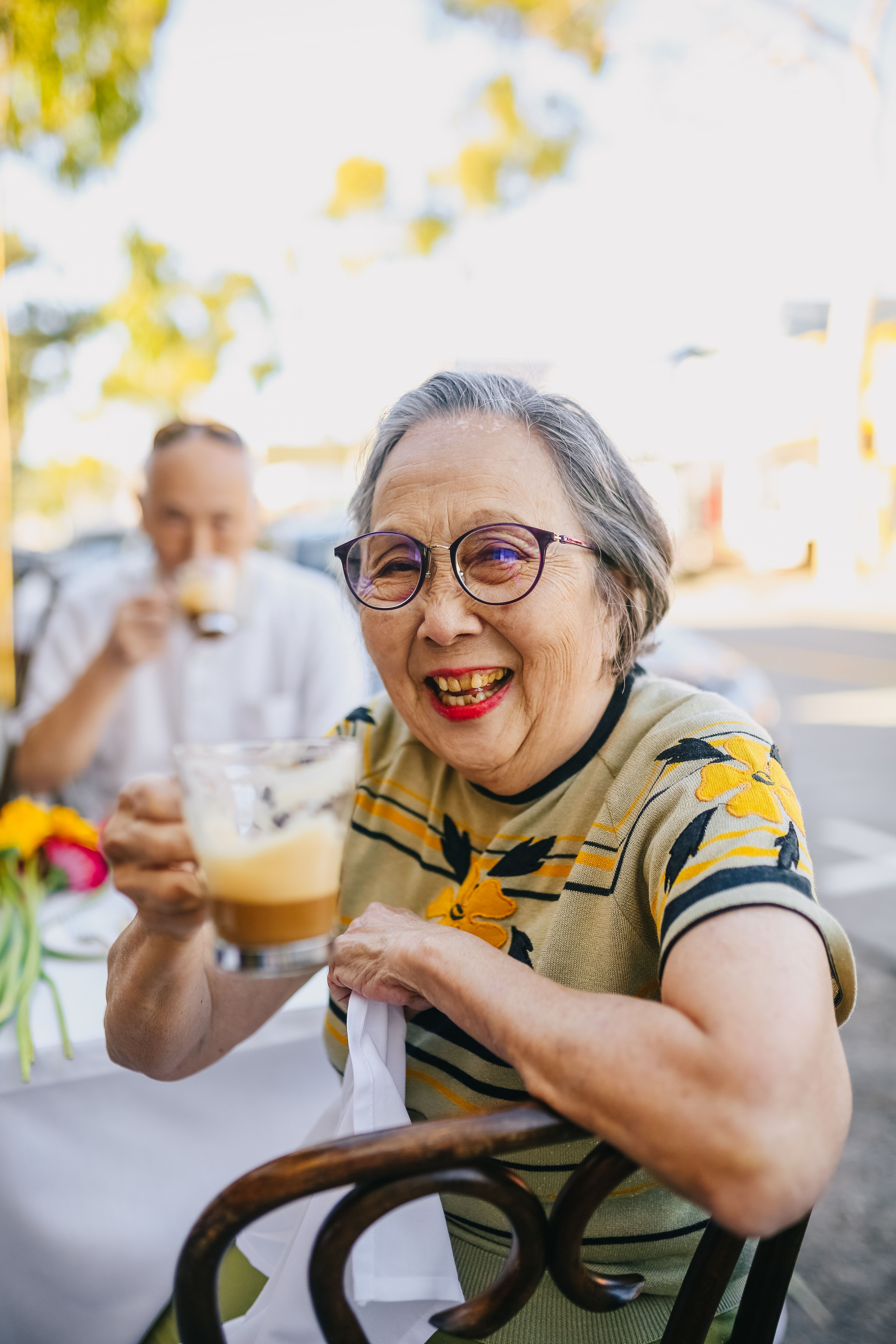 Smiling elderly woman in glasses holding a cup of frothy coffee at an outdoor cafe.