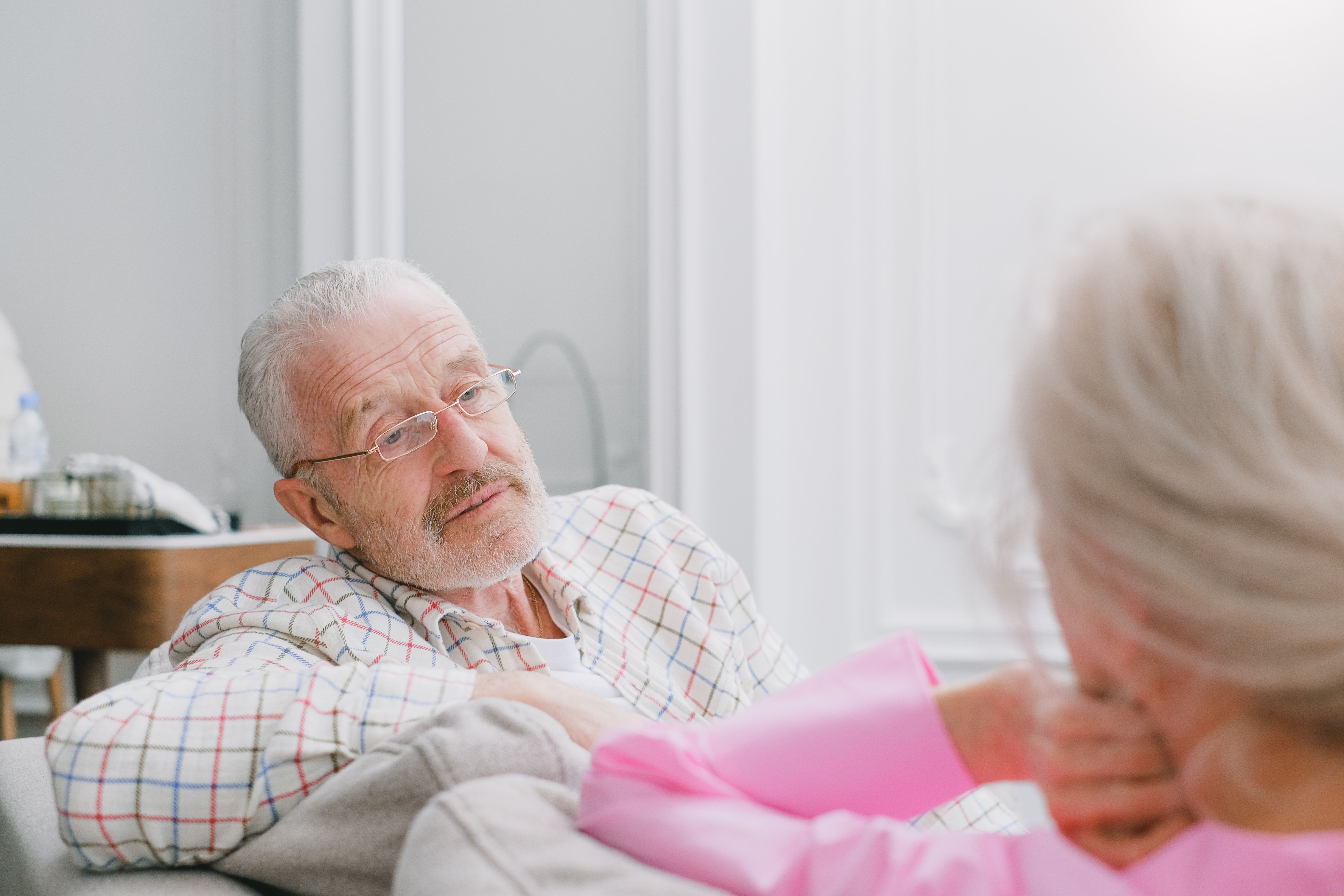 Elderly man with glasses listens intently to an elderly woman in a pink shirt at home.