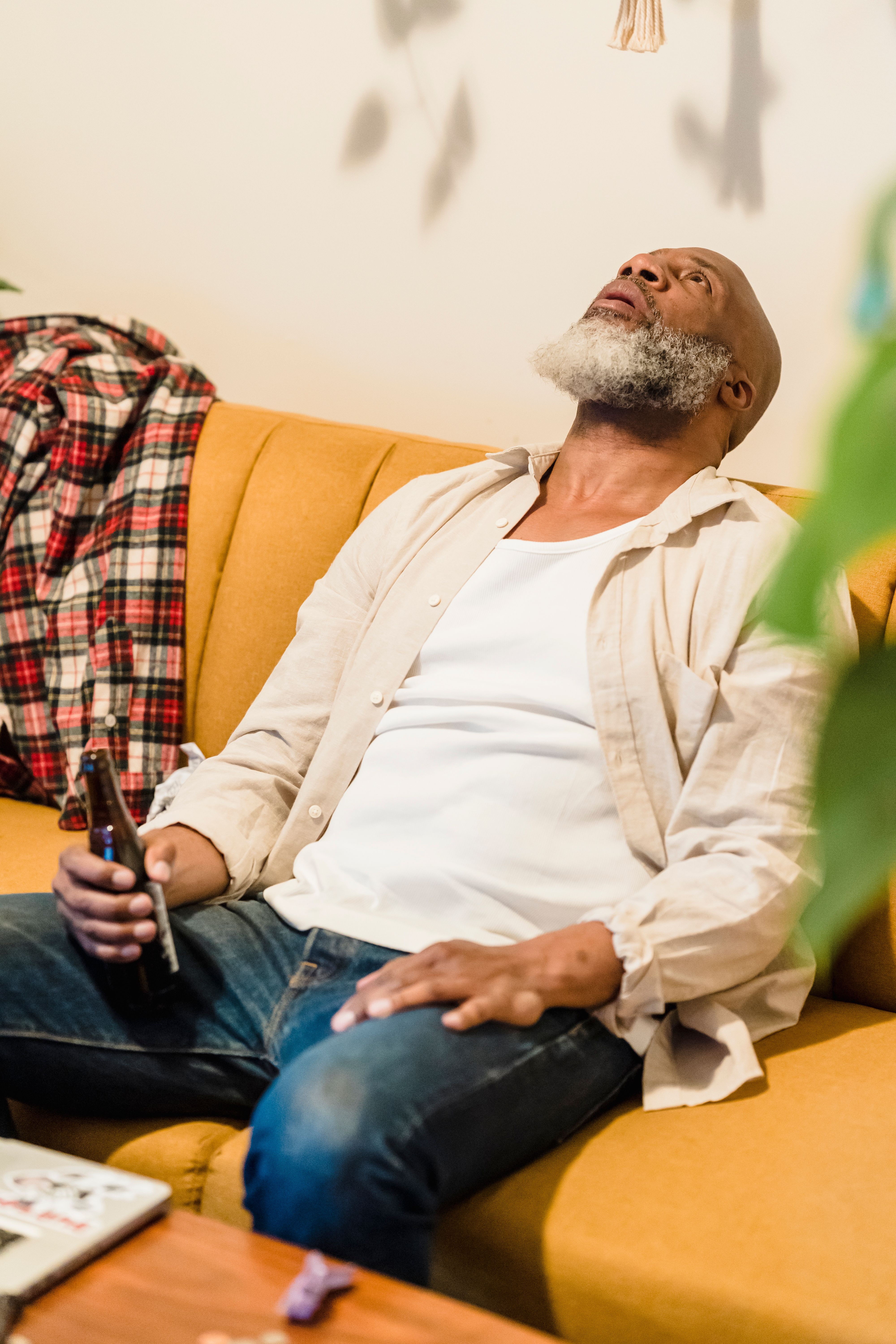 Man with gray beard sitting on a yellow couch looking upward and holding a bottle in his hand.