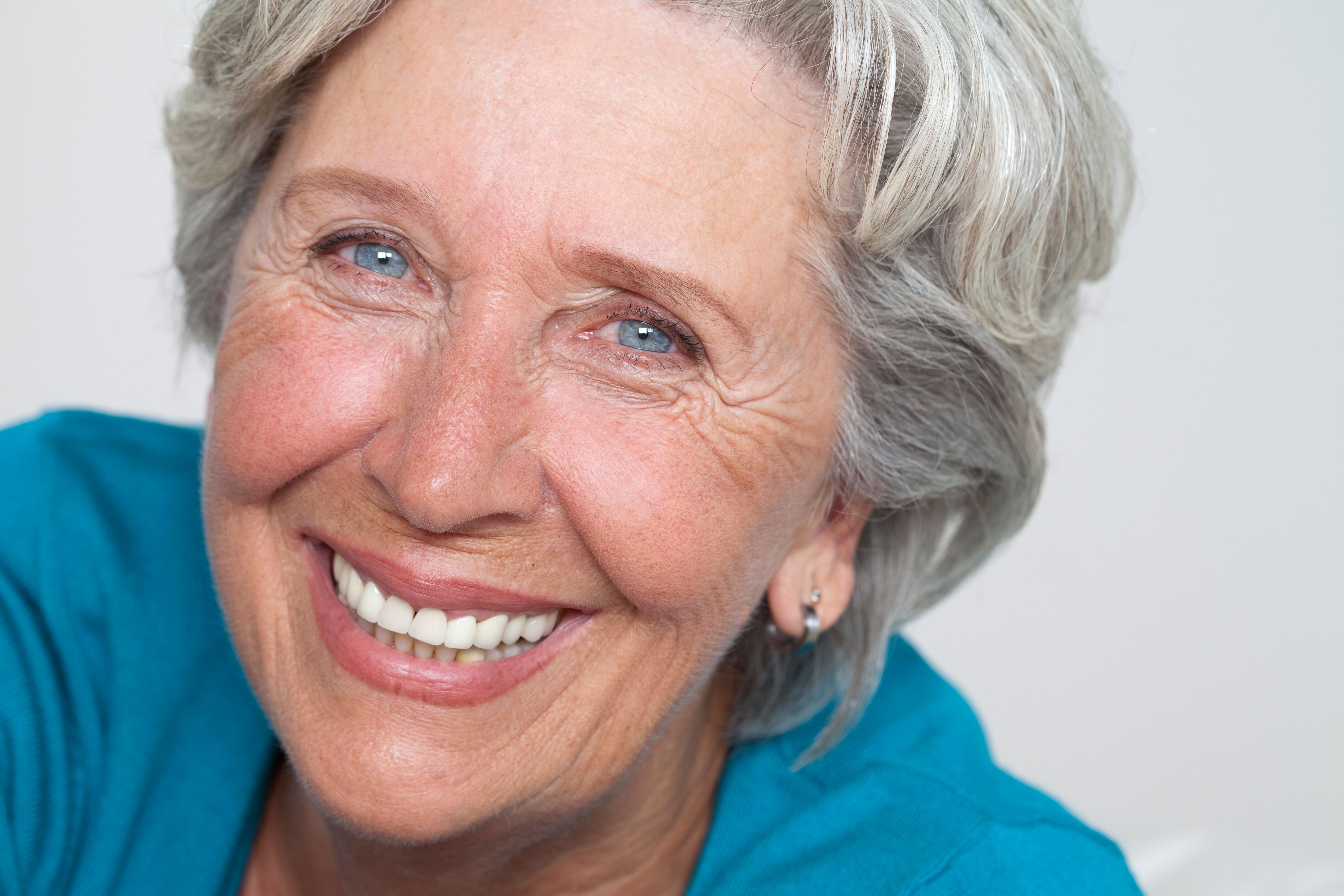 Close-up of an elderly woman with gray hair smiling, showing white teeth and wearing a blue top.