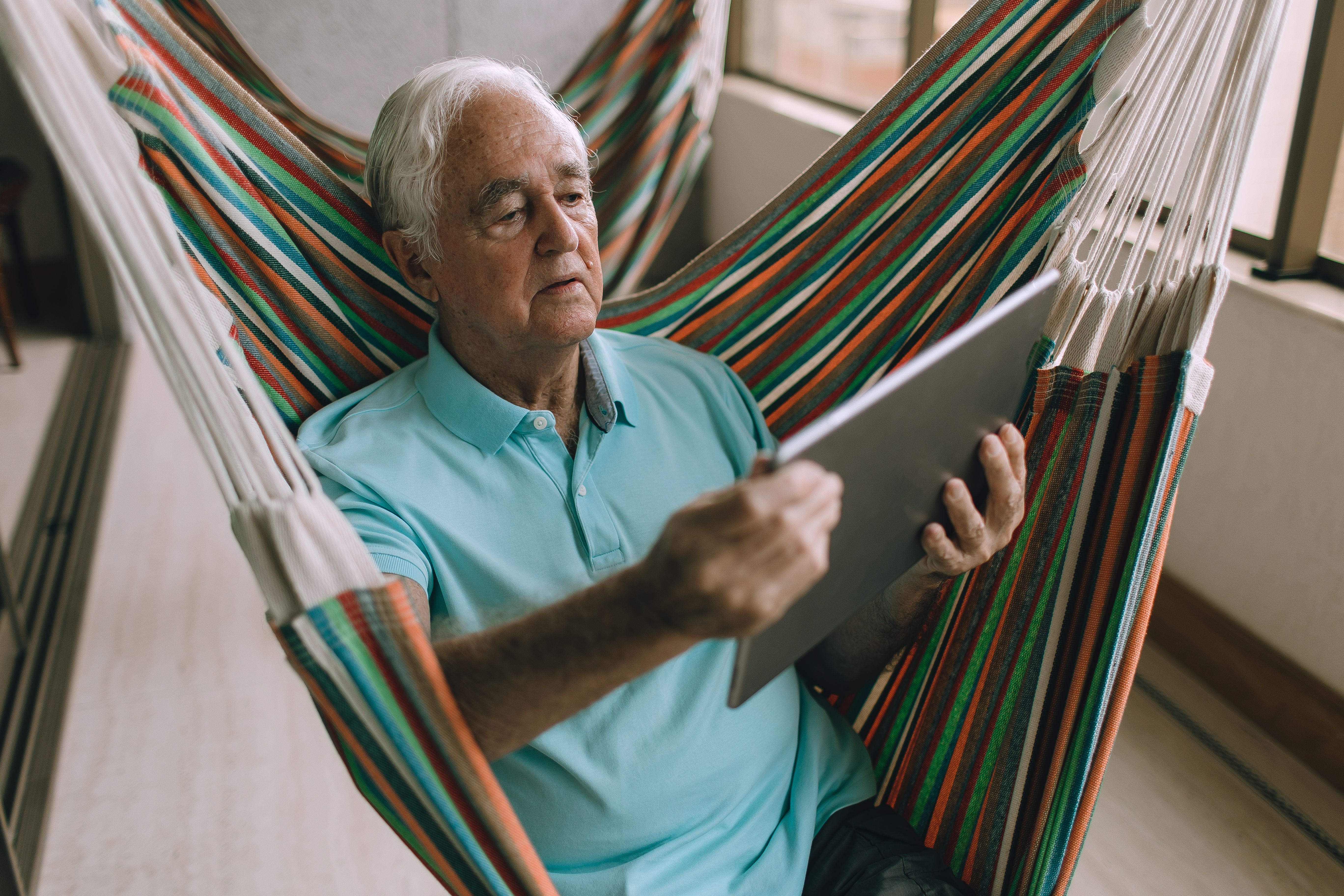 Elderly man in a blue polo shirt reading or using a tablet while sitting in a colorful striped hammock indoors.