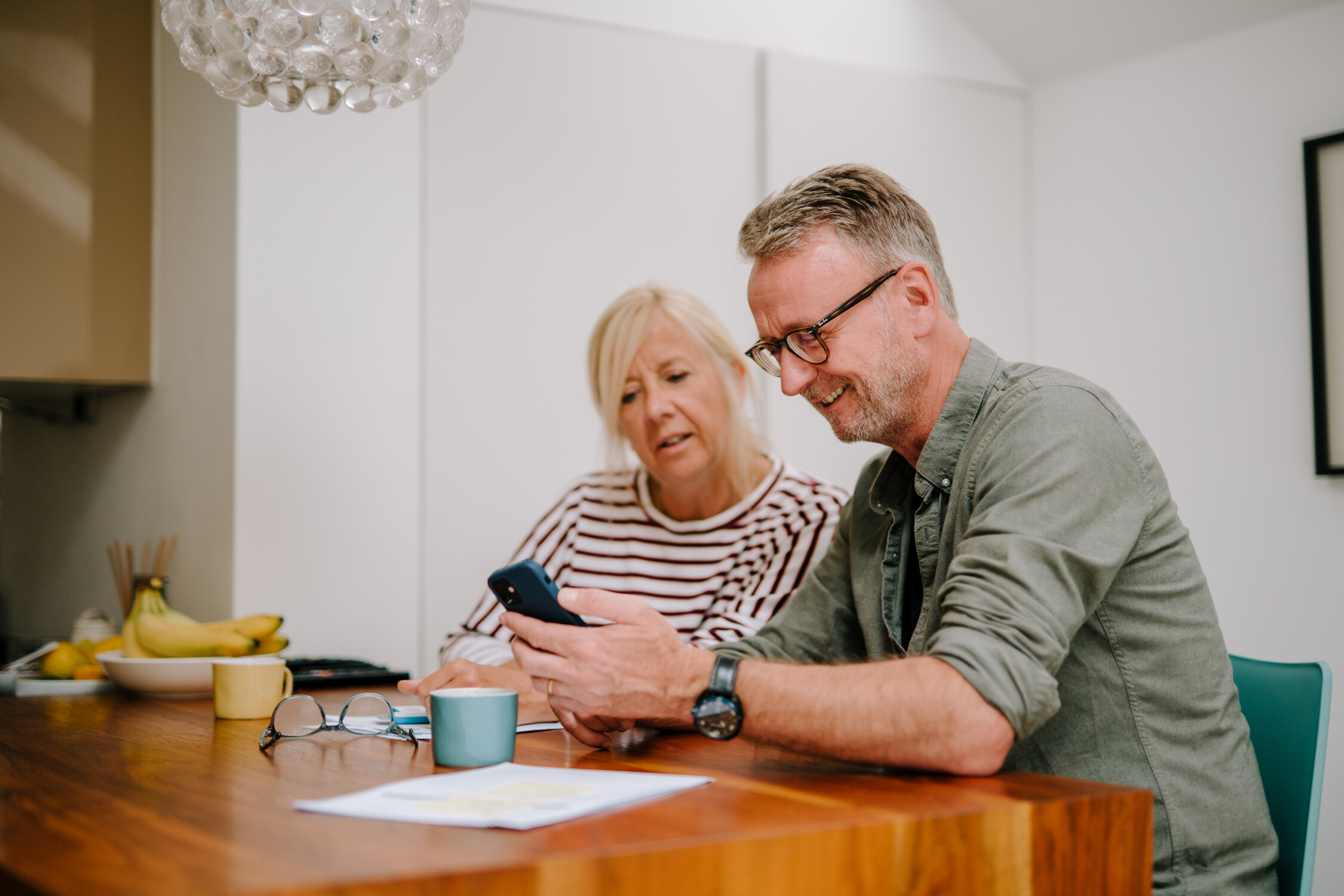 Smiling man shows a smartphone to a woman as they sit at a wooden table in a kitchen.