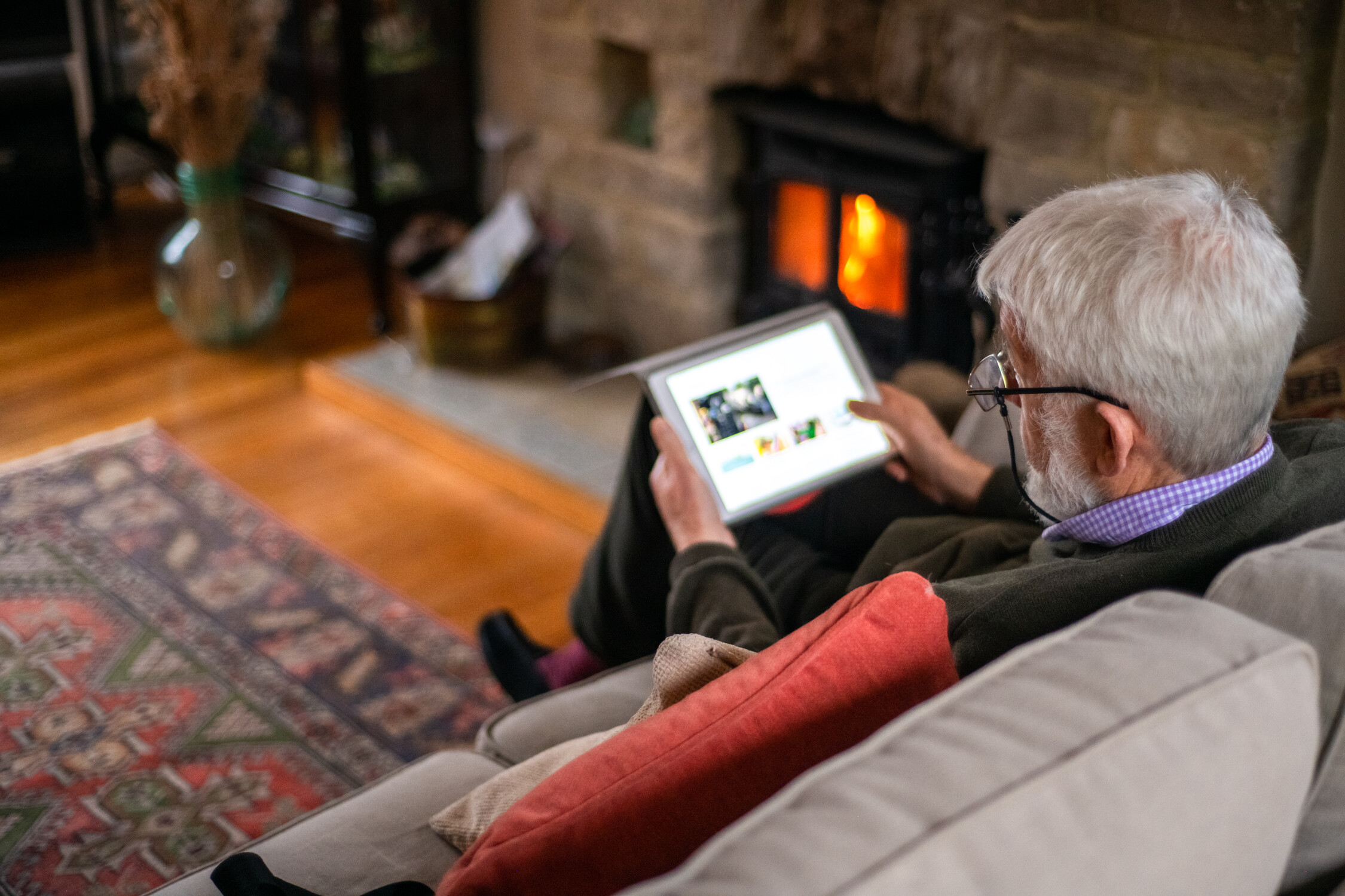 Older man with glasses sitting on couch by lit fireplace using a tablet device.