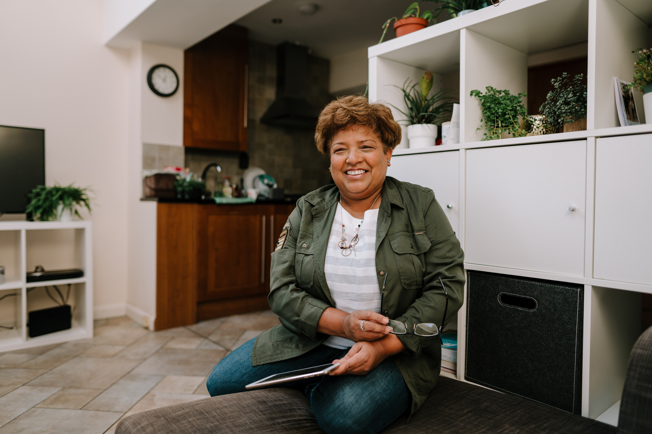 Smiling woman sitting indoors holding glasses and a tablet with plants and kitchen in background.