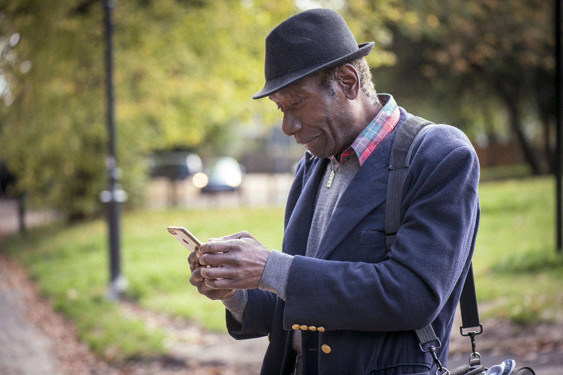 Man in a blue coat and hat smiling while looking at a smartphone outdoors in a park.