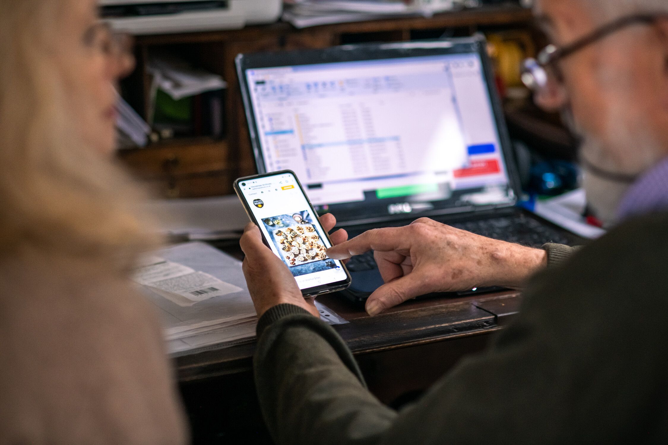 Older man showing an image on his smartphone to a woman, with a laptop in the background.