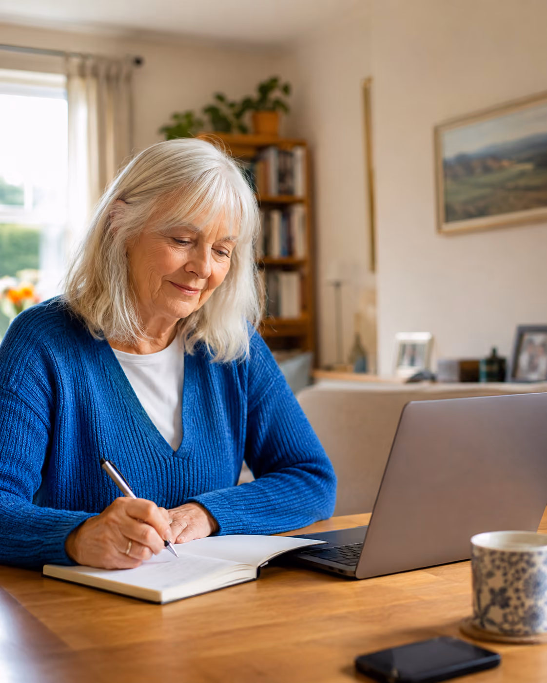 Elderly woman in blue sweater writing in a notebook at a table with a laptop and a cup.