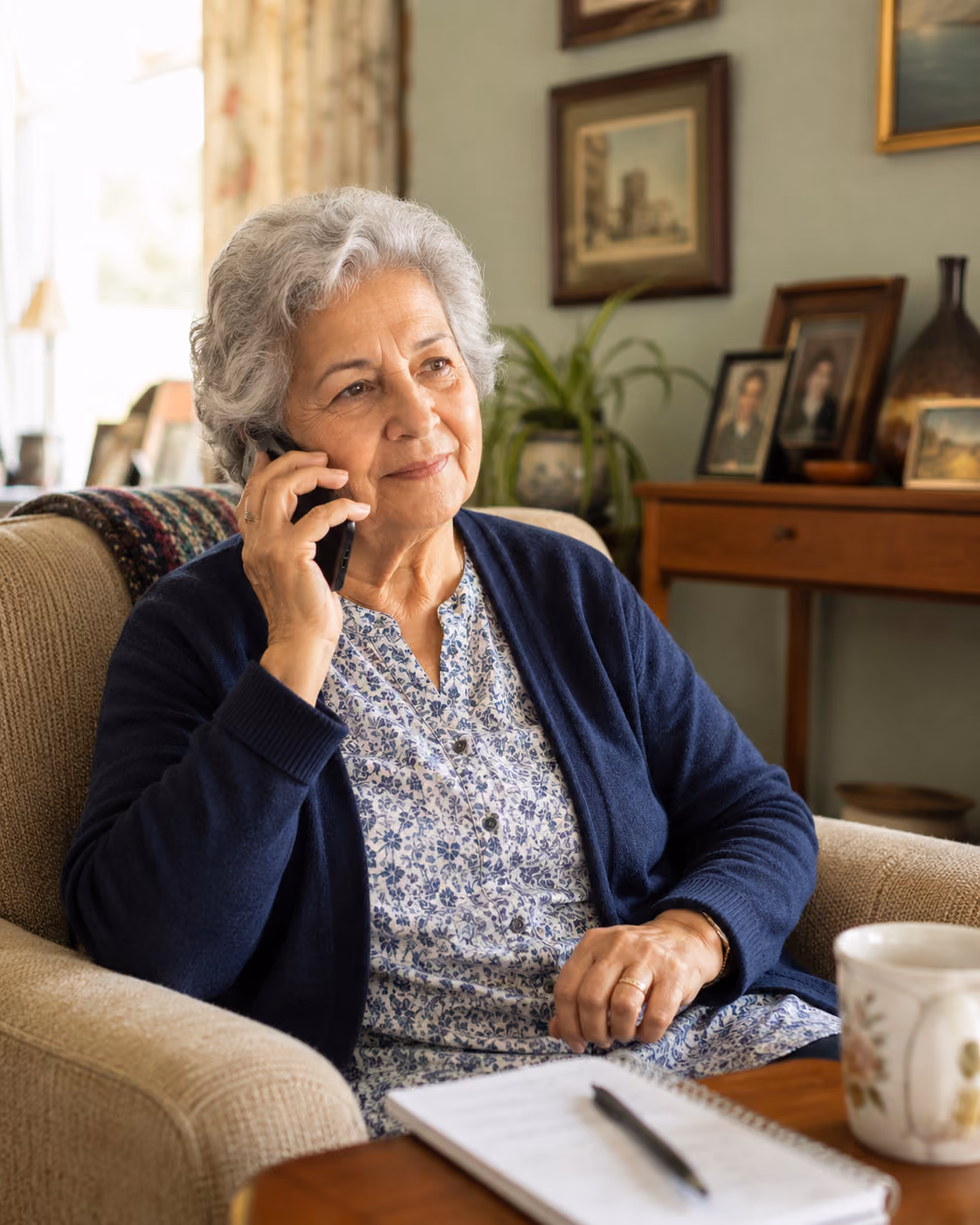 Older woman in navy cardigan sitting on couch, talking on phone with notebook and pen on table.