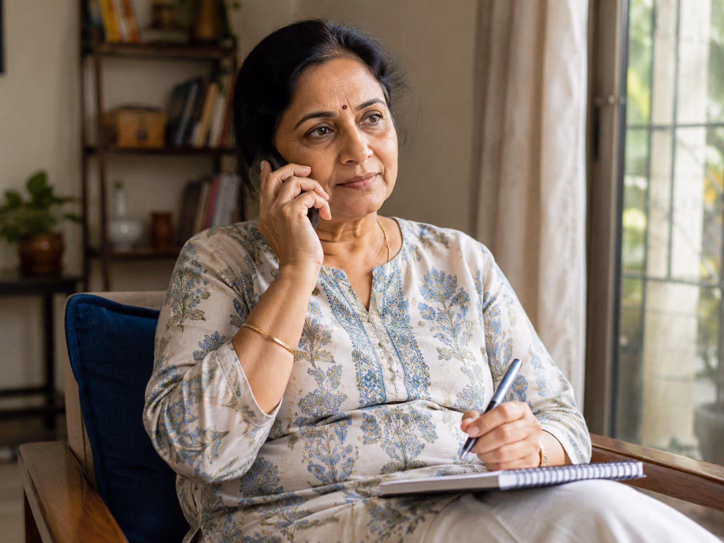 Middle-aged woman in floral blouse on phone, holding pen and notebook, sitting in armchair indoors.