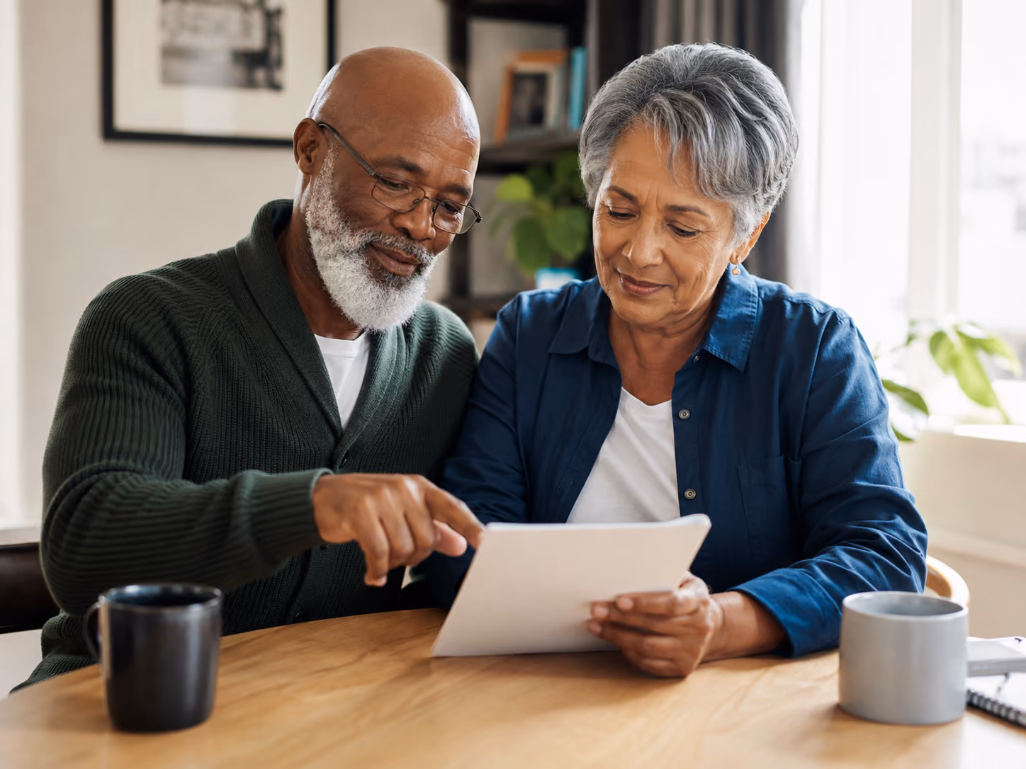 Senior couple sitting at a table reviewing a document together with coffee mugs nearby.