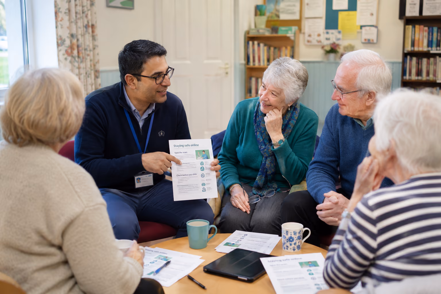 Man explains staying safe online to a group of elderly people in a cozy room with papers and mugs on table.