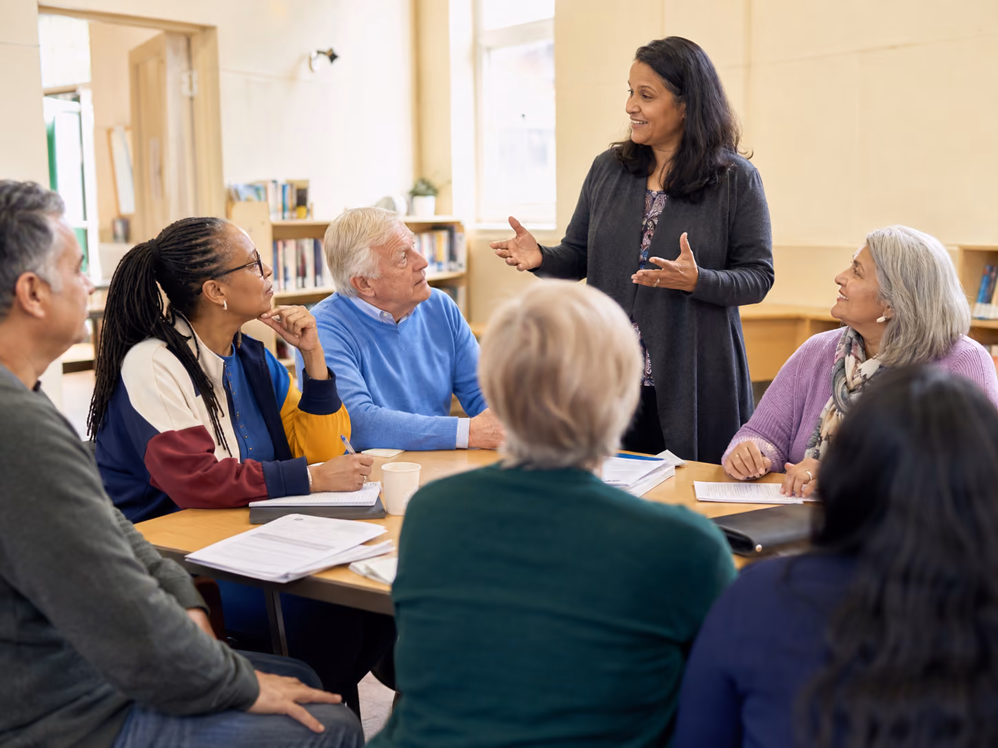 Woman standing and speaking to a diverse group seated around a table with papers in a discussion.