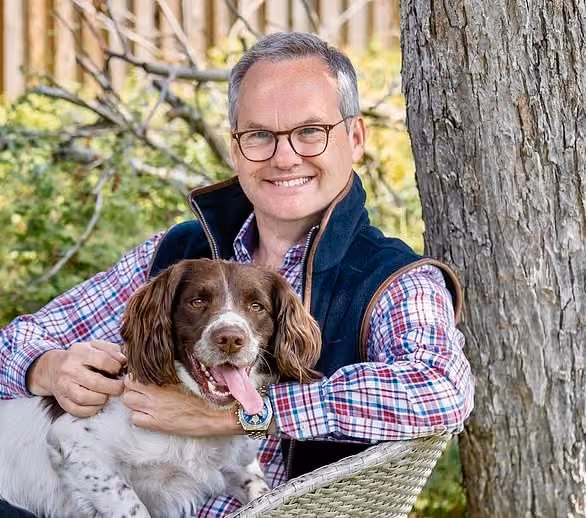 Smiling man with glasses sitting outdoors, holding a happy brown and white dog on his lap.
