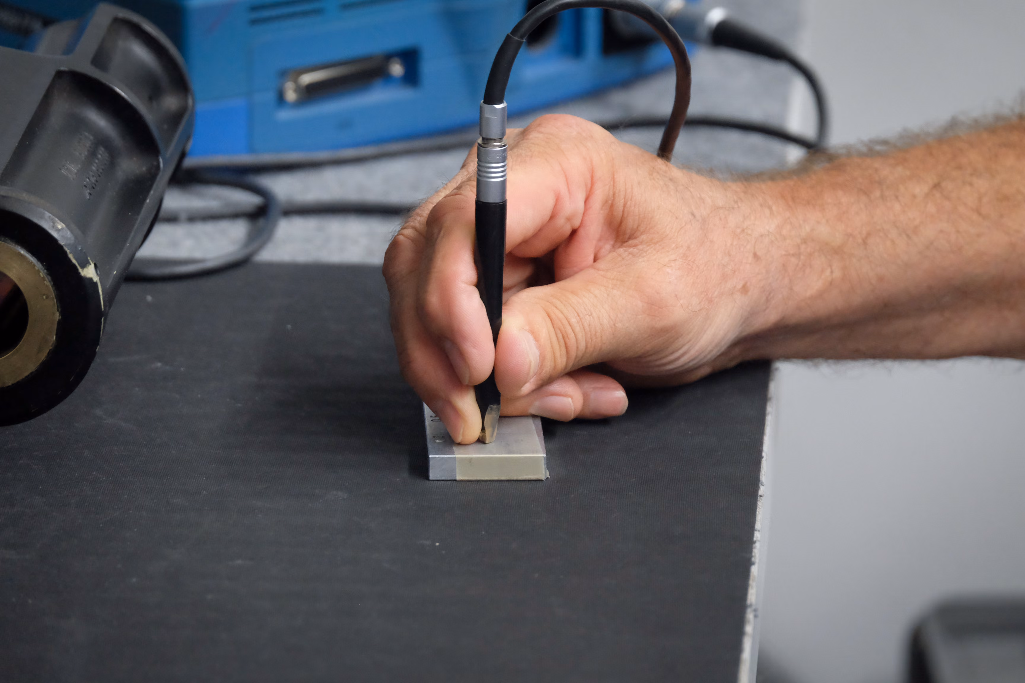 Hand holding a probe touching a metal block on a black surface with equipment in the background.