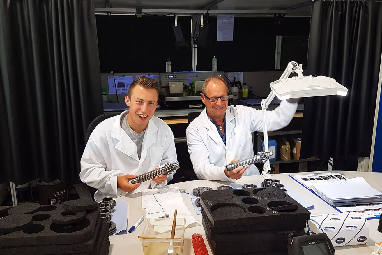 Zwei Männer in weißen Laborkitteln halten Metallteile und lächeln in einer Werkstatt.