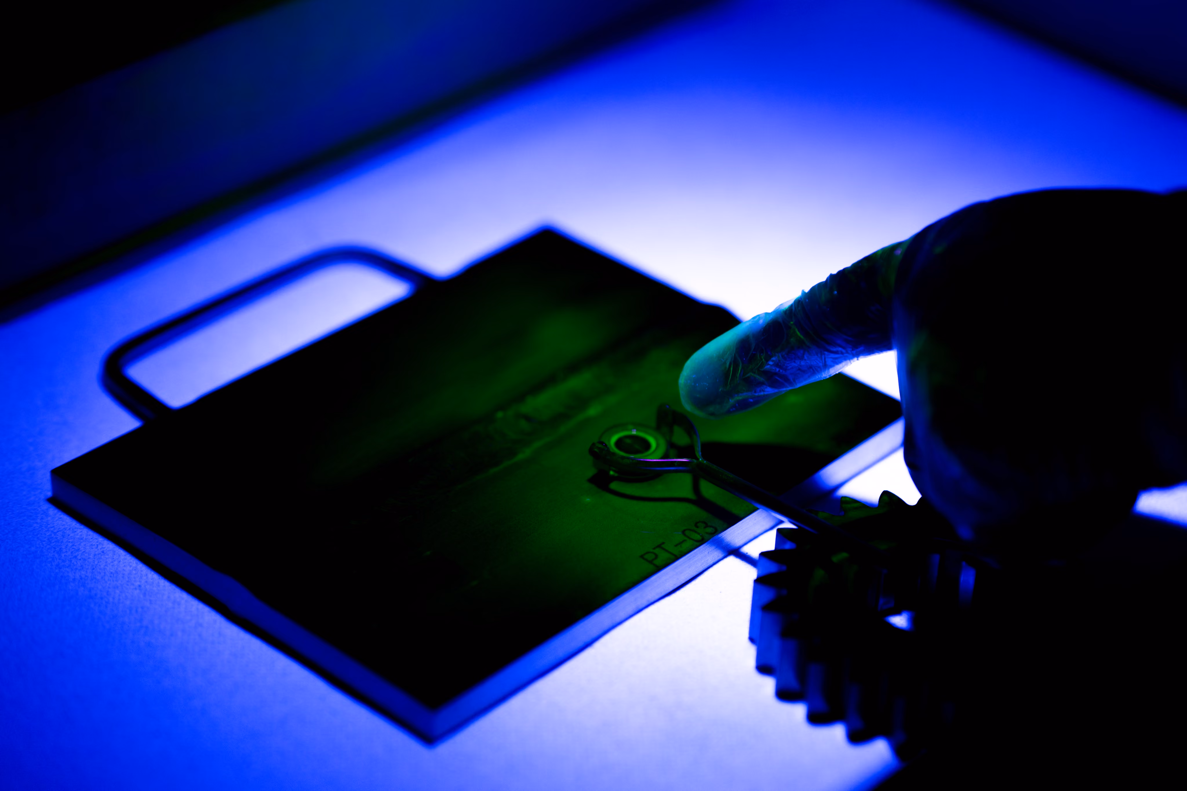Hand with glove holding metal piece near a gear on a blue-lit surface in a workshop setting.