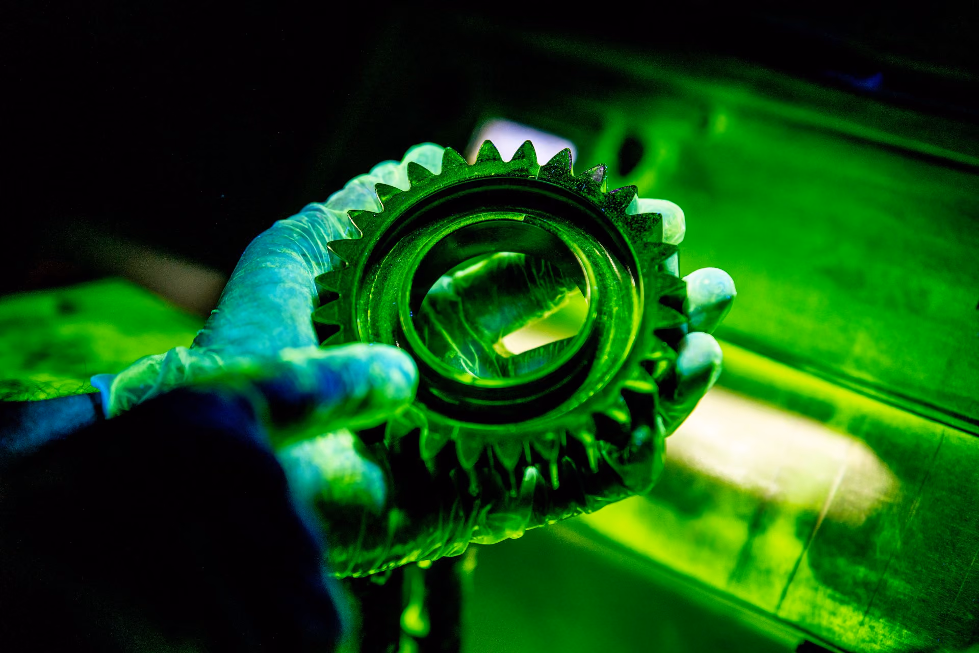 Hand with glove holding a glowing green gear under UV light in a dark setting.