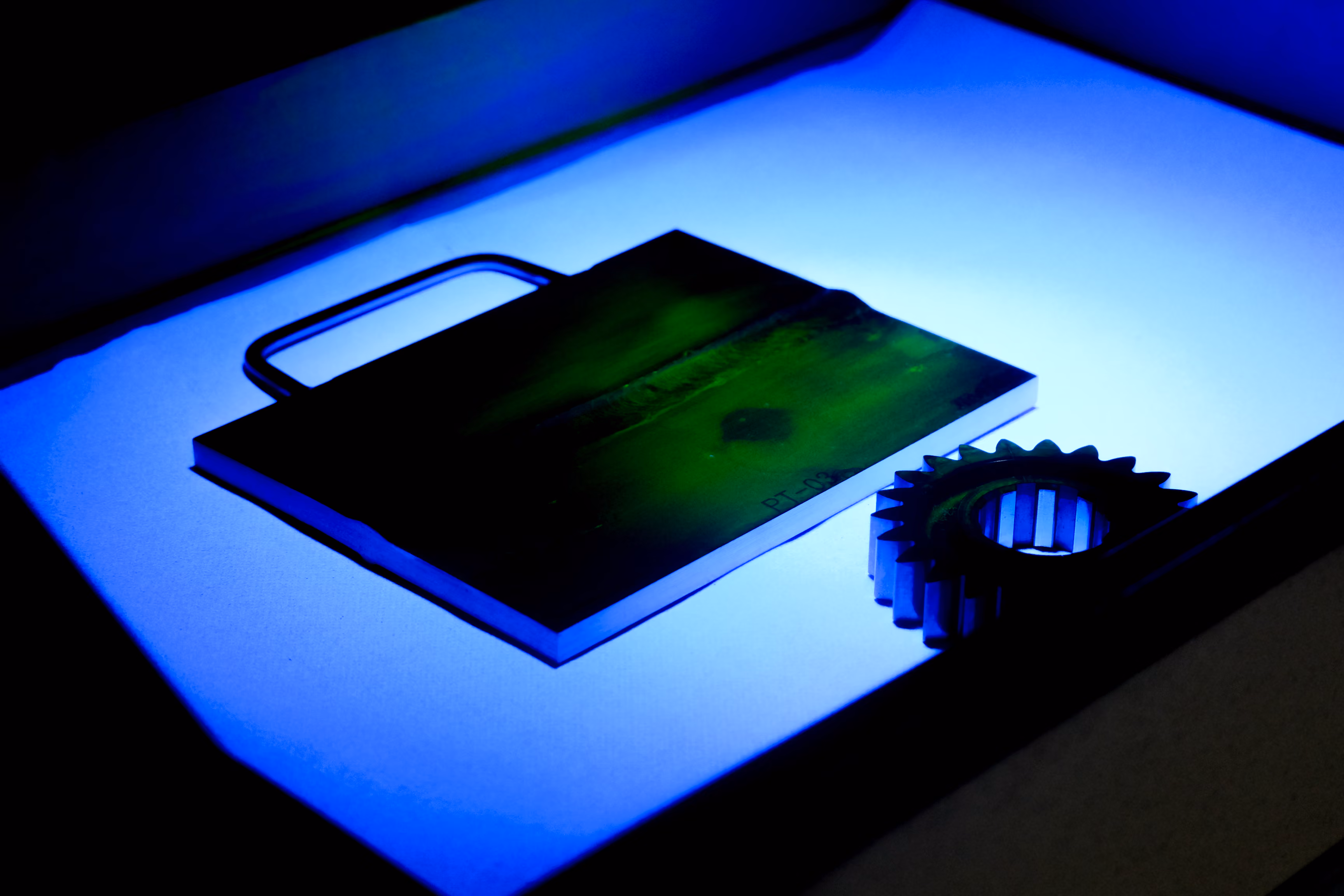 Metallprobe und Zahnrad unter blauem UV-Licht auf weißer Oberfläche.