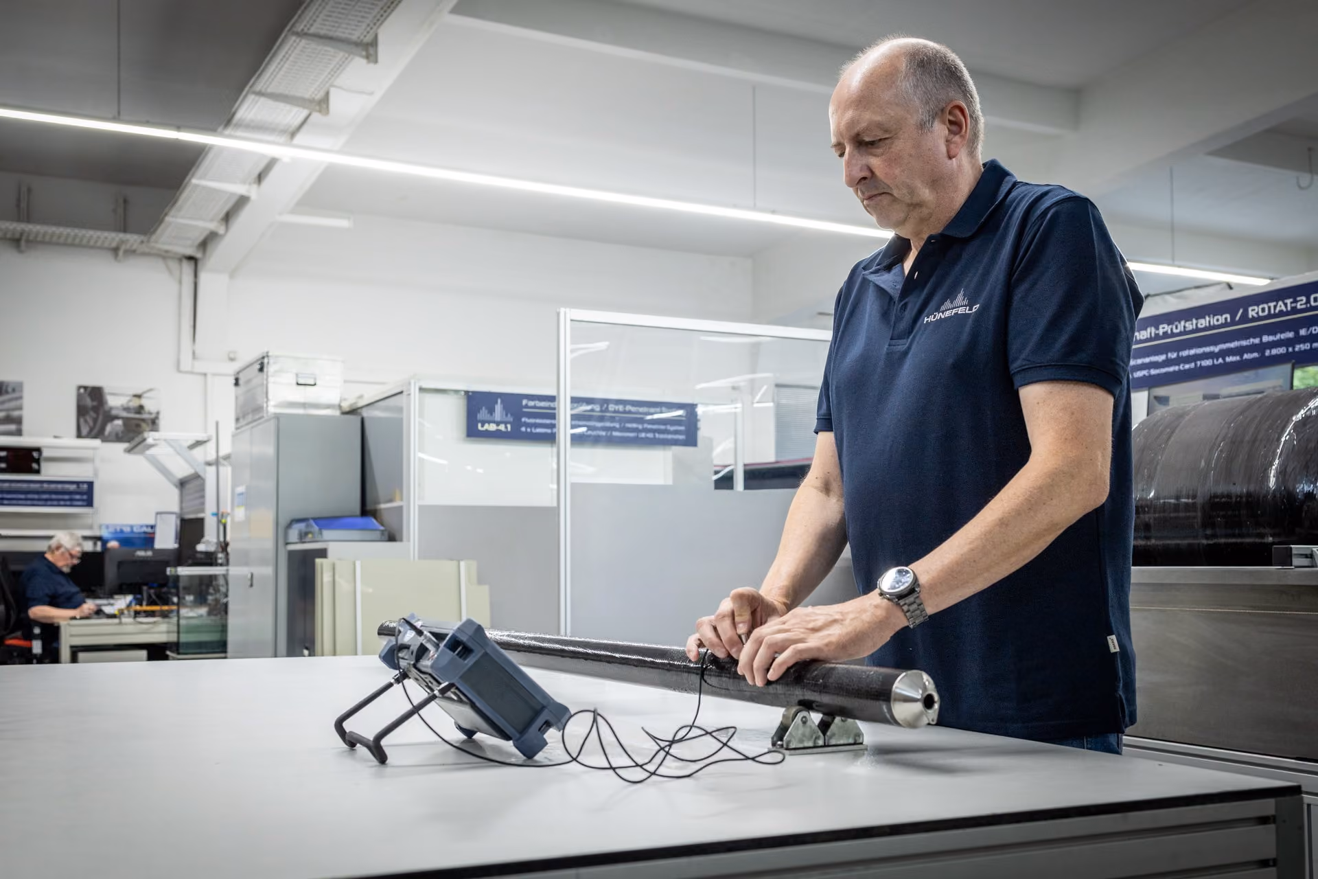Techniker prüft ein langes Carbonrohr mit Messgerät auf einem Labortisch in einer Werkstatt.