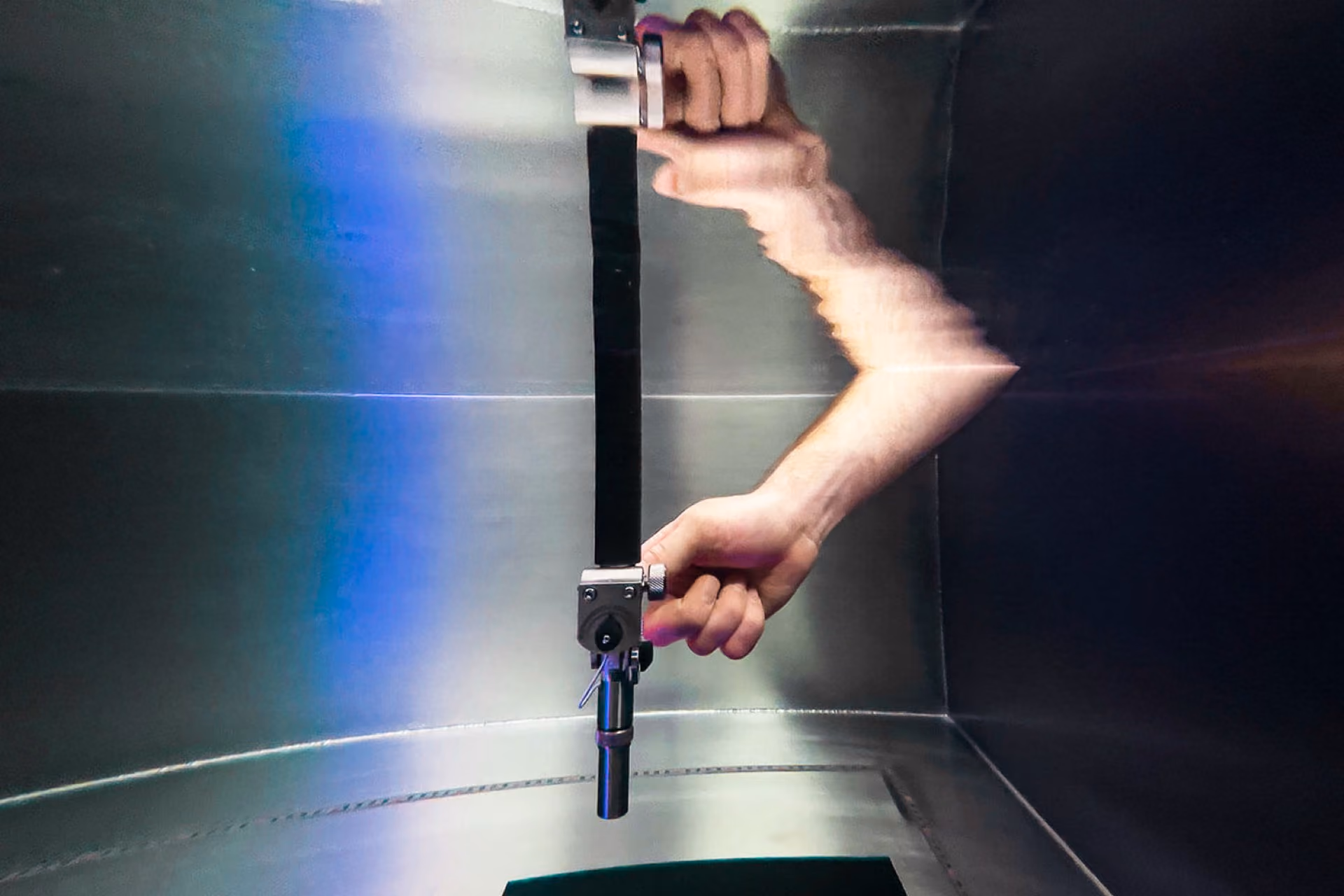 Hand holding a black rod with a metallic clamp inside a reflective metal chamber with blue light.