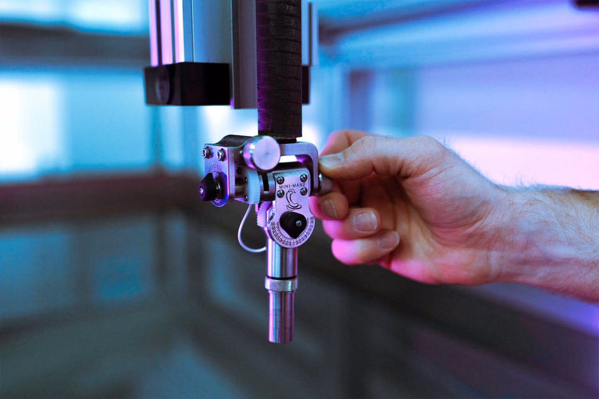 Hand adjusts a metallic mechanical device labeled MINI-MAN2 with purple-blue ambient light.