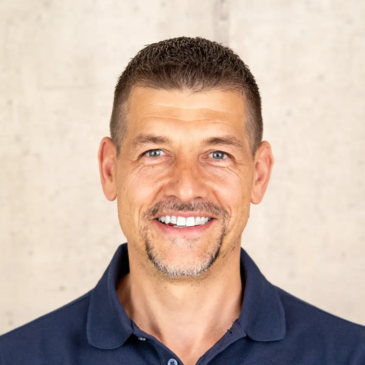 Lächelnder Mann mit kurzen braunen Haaren und blauem Polohemd vor beigem Hintergrund.