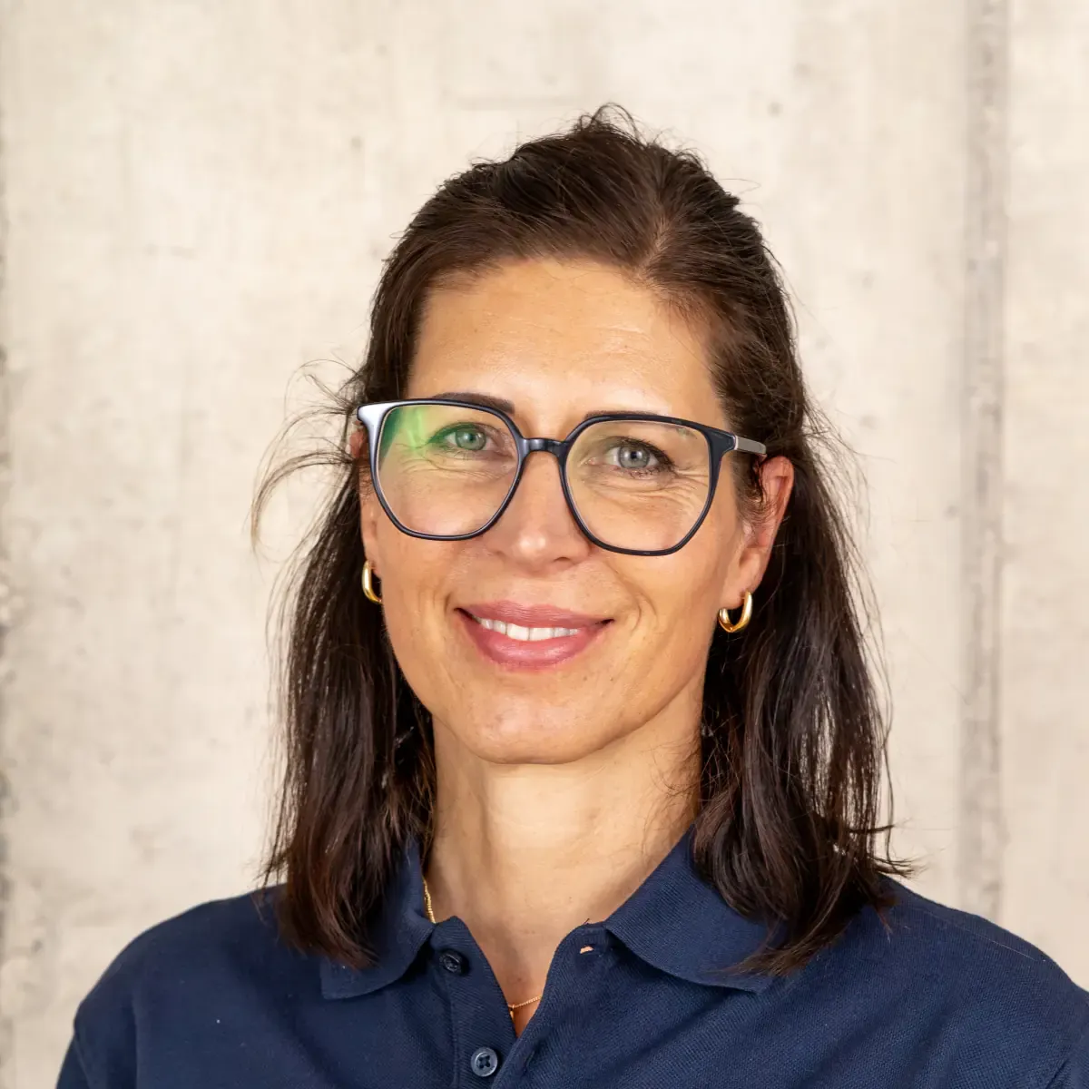 Frau mit braunen Haaren, Brille und blauer Bluse vor beigem Hintergrund lächelt freundlich.