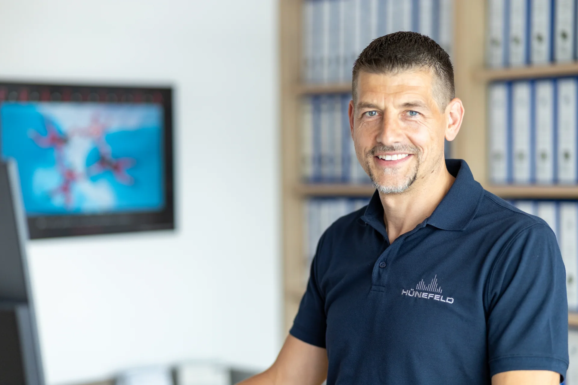 Mann in blauem HÜNEFELD-Poloshirt lächelt in einem Büro mit Regalen voller Ordner.