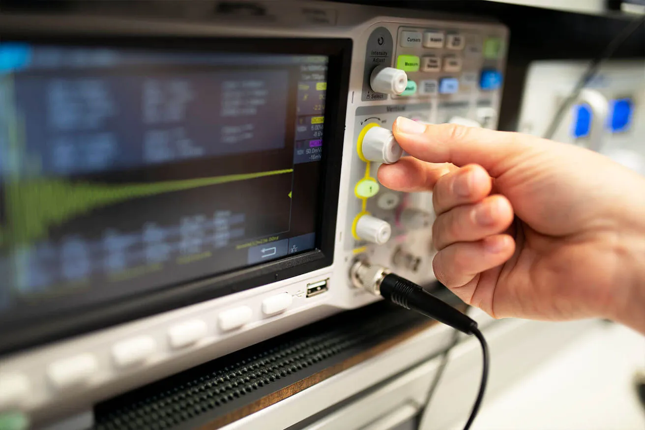 Hand adjusting a control knob on an oscilloscope displaying a waveform on screen in a lab.