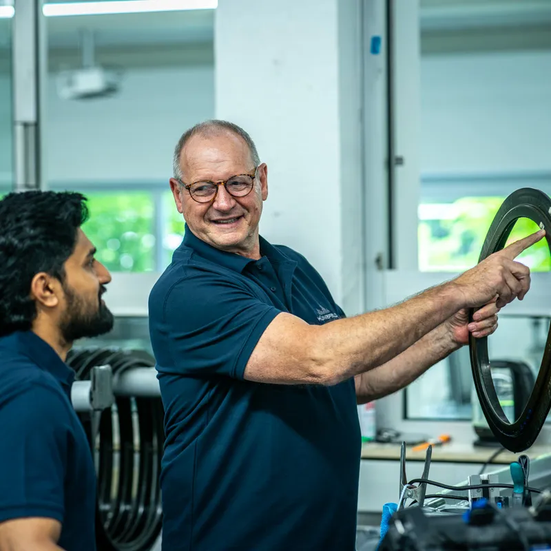 Zwei Männer in blauen Poloshirts besprechen ein rundes Metallteil in einer Werkstatt.