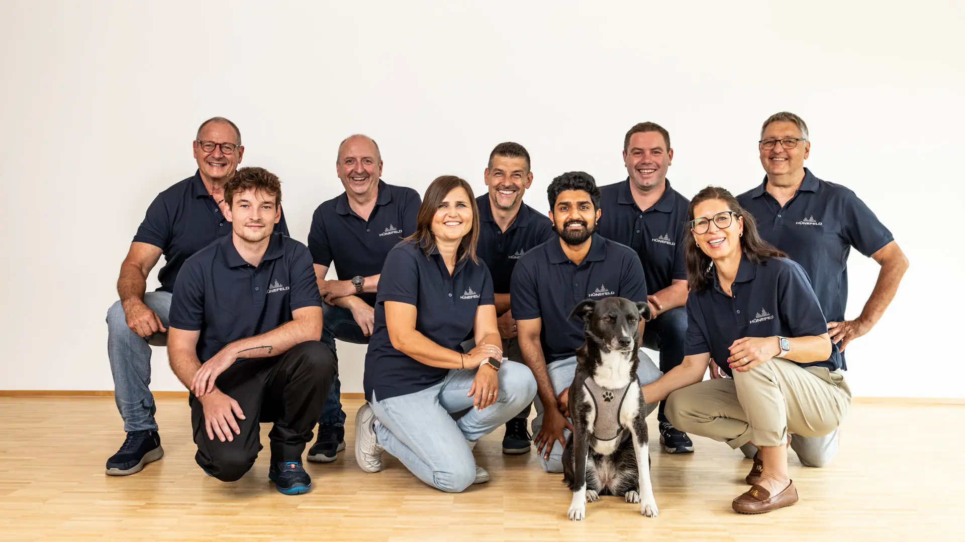 Neun lächelnde Menschen in blauen Poloshirts mit Hund sitzen und knien auf hellem Holzboden.