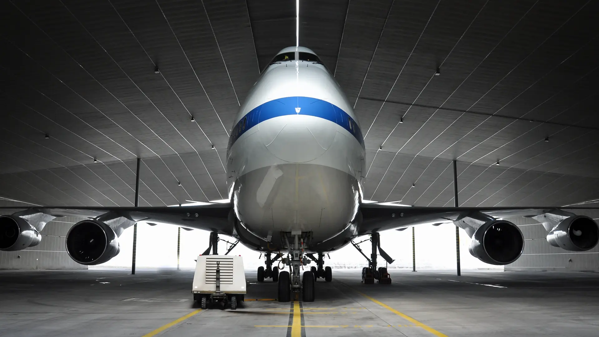 Front view of a large airplane parked inside a spacious hangar with lights above.