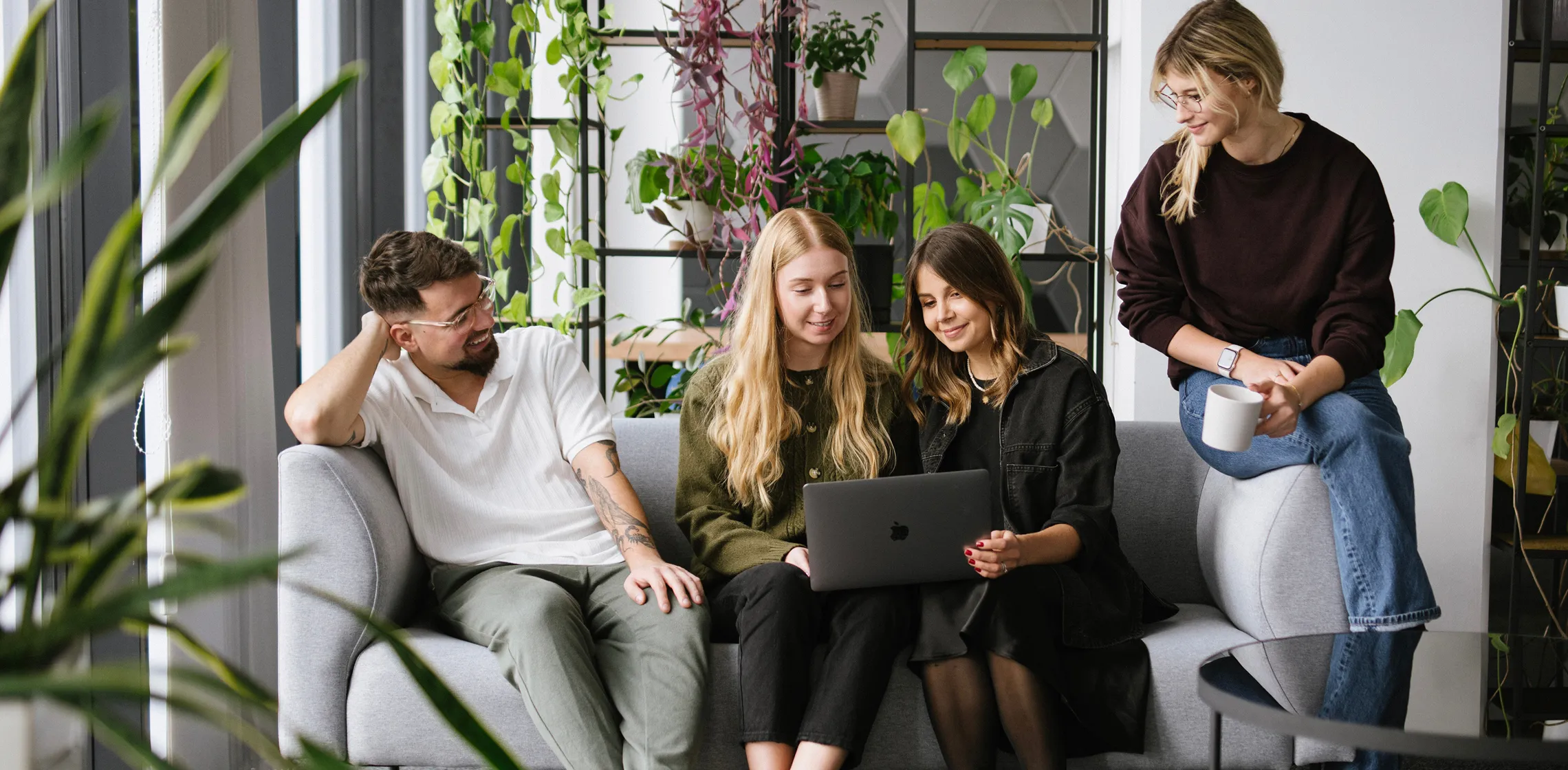 Four young adults sitting and smiling on a couch looking at a laptop in a plant-filled room.