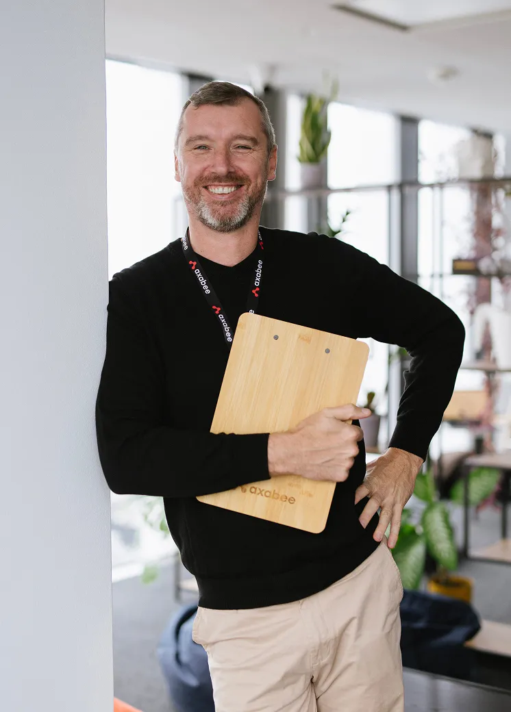 Smiling man in black sweater and beige pants holding a wooden clipboard in an office setting.