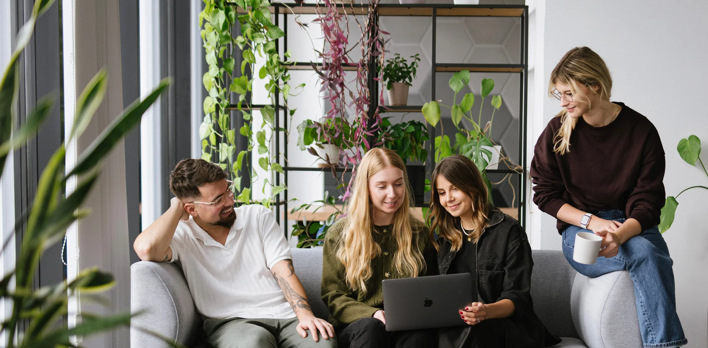 Four young adults sitting on a couch, two looking at a laptop, surrounded by indoor green plants.