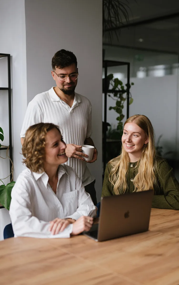 Three coworkers smiling and talking around a laptop at a wooden table in an office.