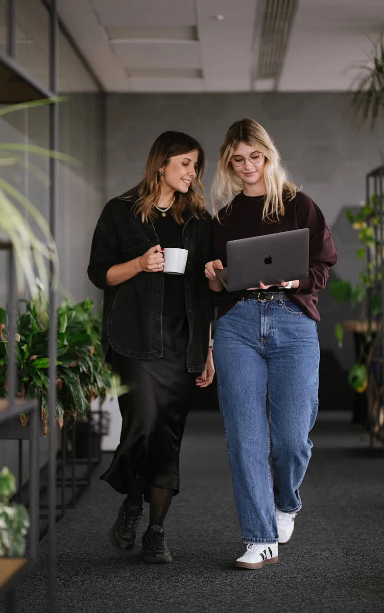 Two women walking in an office, one holding a laptop and the other a white mug with plants around.