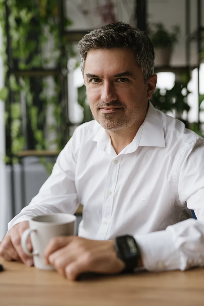 Man in white shirt holding a cup, sitting at a wooden table with plants in the background.