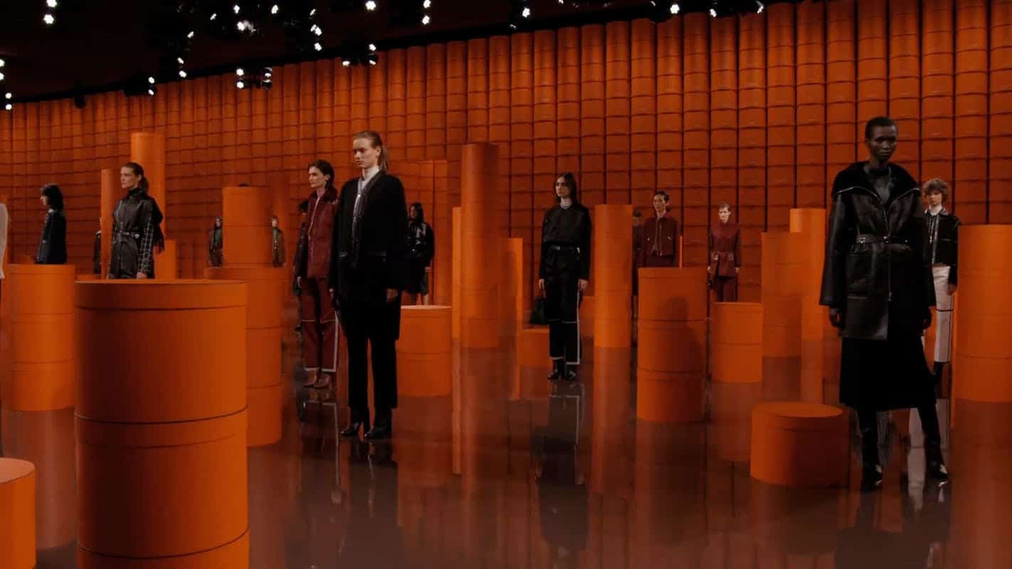 Models stand among orange pedestal columns on a mirrored floor at the Hermès show